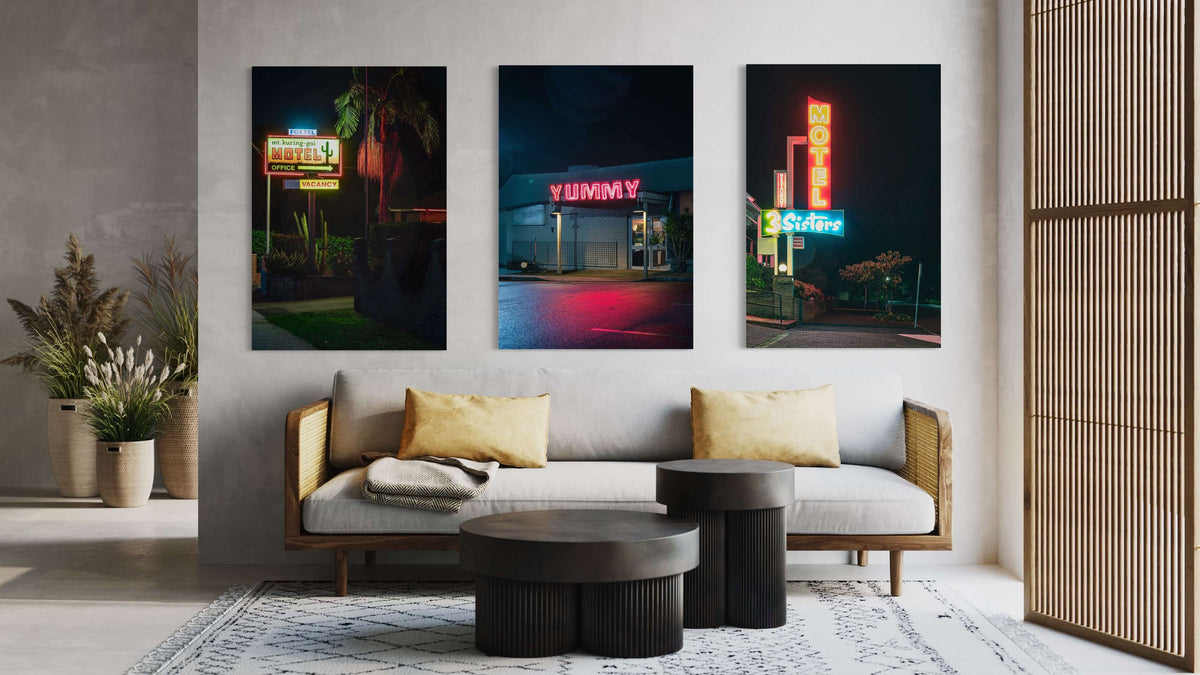 Three Lost Collective neon-sign prints hung above a timber-framed sofa in a styled living room. Left: a motel office sign at night. Centre: a YUMMY diner sign in red neon. Right: a 3 Sisters motel sign.