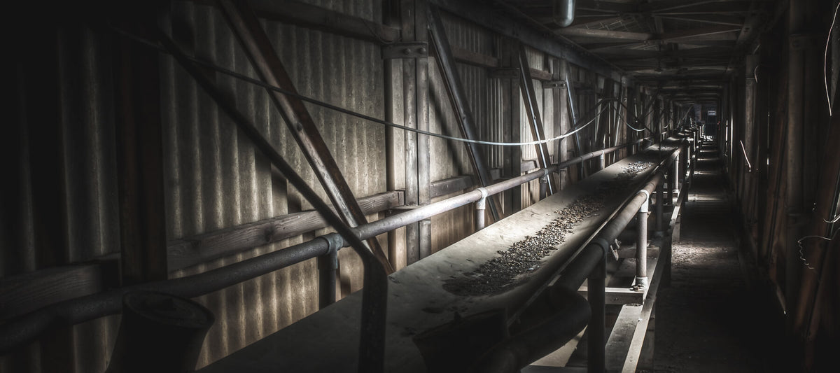 A long pipe-and-conveyor corridor inside the Bradmill mill, lined with corrugated metal walls and diagonal timber bracing, lit by a thin band of light from the far end.