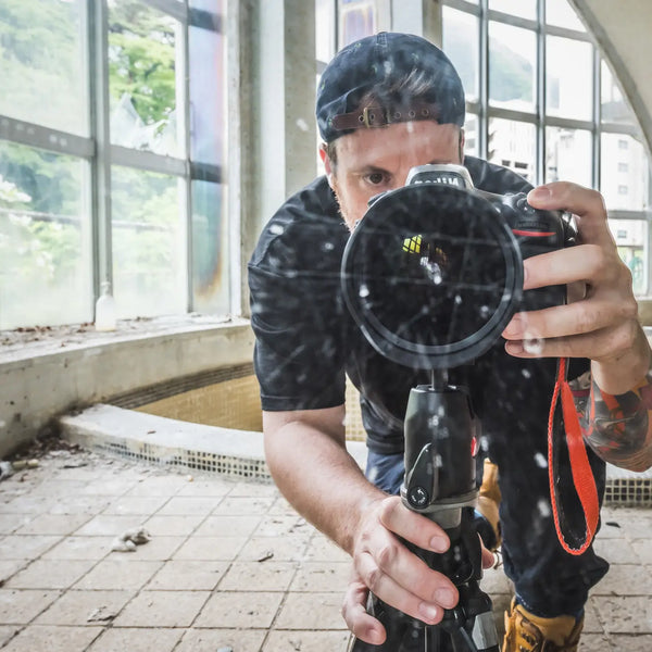 Brett Patman crouched behind a tripod-mounted camera at Kinugawa Kan, lens pointed forward, arched windows and the Kinugawa valley visible behind him.