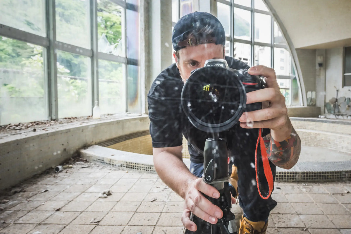 Brett Patman crouched behind a tripod-mounted camera at Kinugawa Kan, lens pointed forward, arched windows and the Kinugawa valley visible behind him.