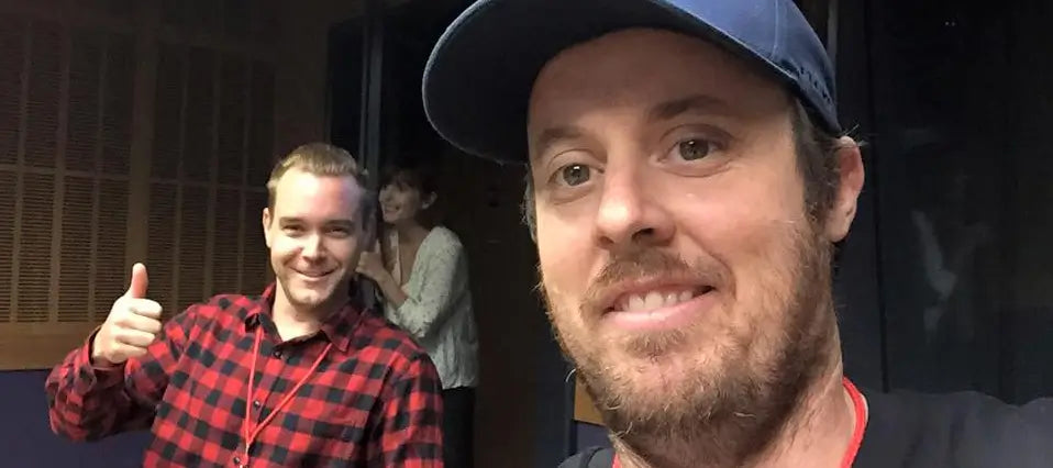 Brett Patman in a navy cap in the foreground, with Tim Frawley in a red and black flannel shirt behind him giving a thumbs up in the ABC Radio National studio. A third person partially visible in the doorway.