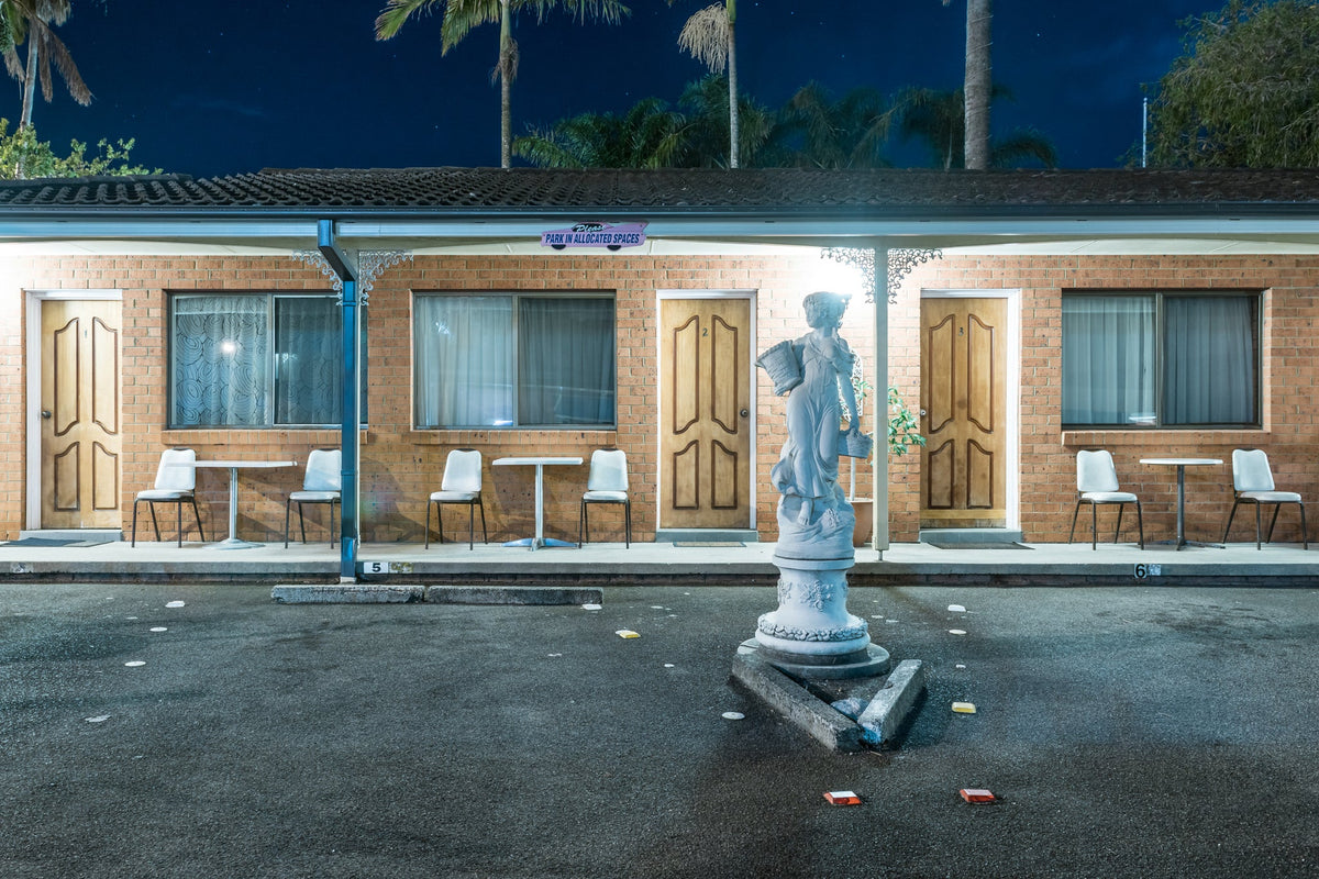 A brick motel at night, a classical statue on a pedestal in the carpark, white chairs outside numbered doors, palms behind.