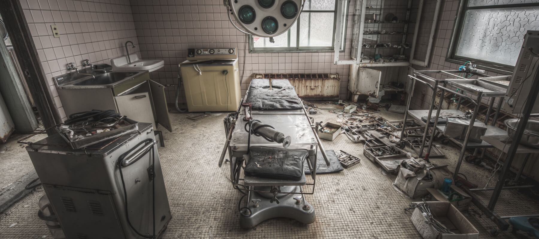 An abandoned operating room at Kuwashima Hospital, with a surgical table, medical equipment, and scattered tools, highlighting the decay and disrepair of the facility.