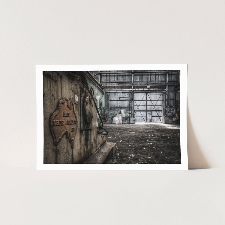 A conveyor drive casing with a 'LOT 23' label affixed, inside a surface building at Awaba Colliery, with bright light pushing through a corrugated metal door at the far end.
