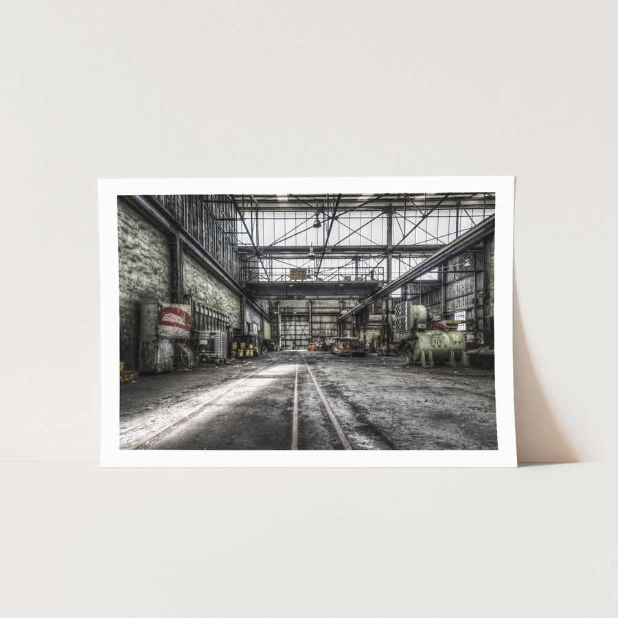 Rusted cylindrical machinery and twin rail tracks on the workshop floor at Awaba Colliery, with a Johnson-Hepburn sign hanging from a gantry crane and grease buckets lining the left wall.
