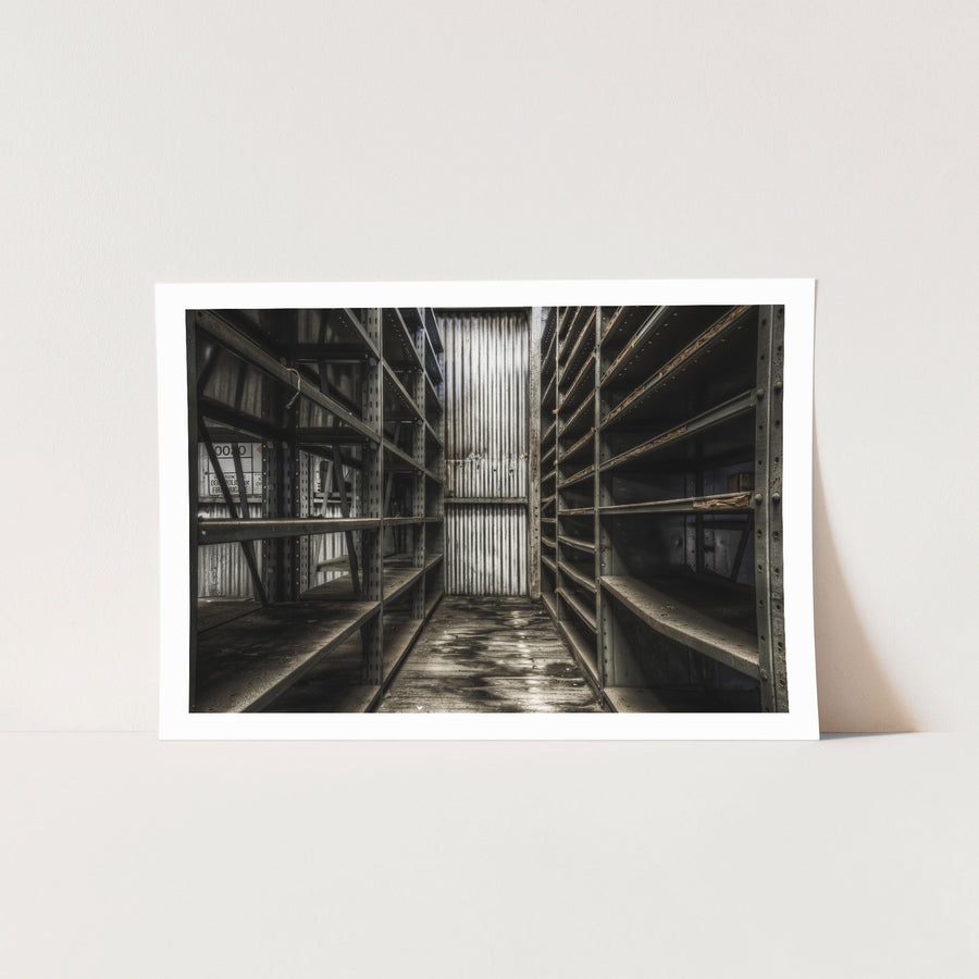 Rust-streaked metal shelving lining a narrow aisle in a derelict storeroom at Awaba Colliery, with warped floorboards and a partially legible sign visible on the left unit.