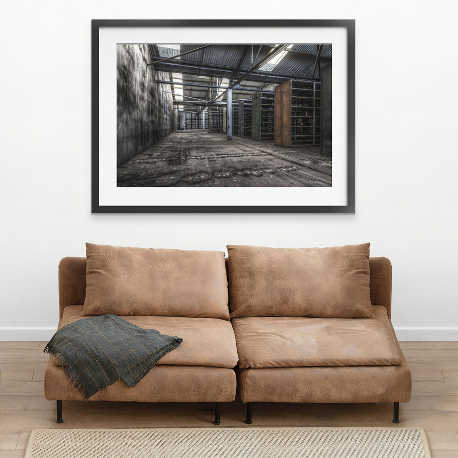 Emptied metal shelving units receding through the upper storeroom at Awaba Colliery, with skylights in a corrugated iron roof casting light across water-stained concrete walls and worn timber floorboards.