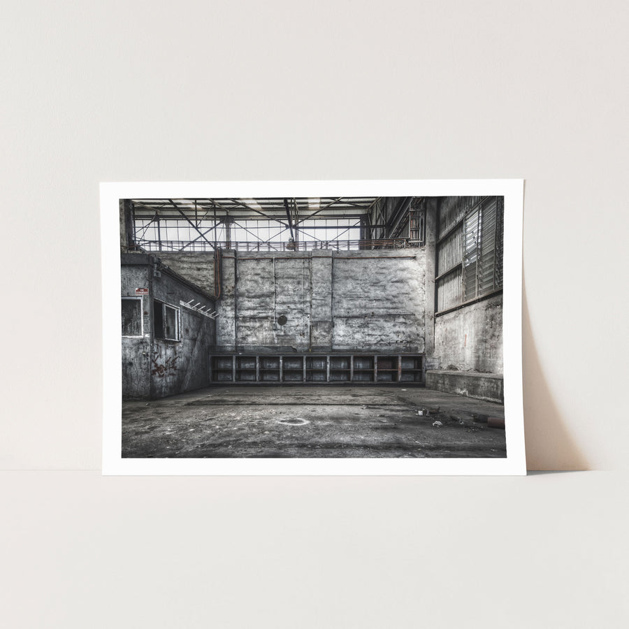 Empty shelving units and a faded fire extinguisher sign on the rendered wall of a workshop bay at Awaba Colliery, with graffiti near two windows and exposed pipes overhead.
