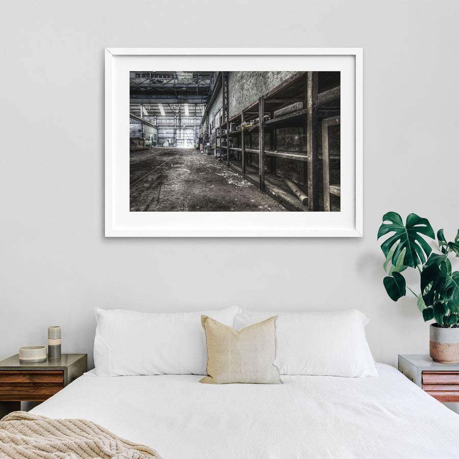 Rusted workshop shelving and faint floor rail tracks inside the colliery workshop at Awaba, with a crane bearing the name "Morison Bearby" visible overhead and a white cup resting on the concrete floor.