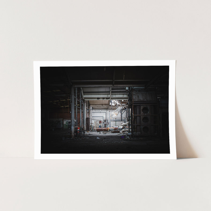 A collapsed sheet-metal roof section hanging across the rear wall of a derelict manufacturing hall at the former Davies Coop / Bradmill factory in Yarraville, with a large rusted cylindrical pipe and scattered debris on the concrete floor.