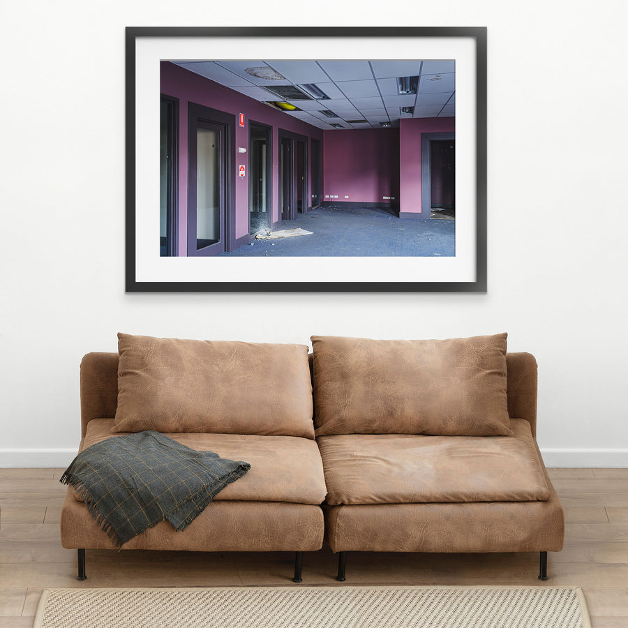 Empty office corridor at the former Bradmill factory in Yarraville, with mauve walls, grey carpet, glazed timber-framed doorways, a damaged suspended ceiling, and a red fire safety sign on the wall.
