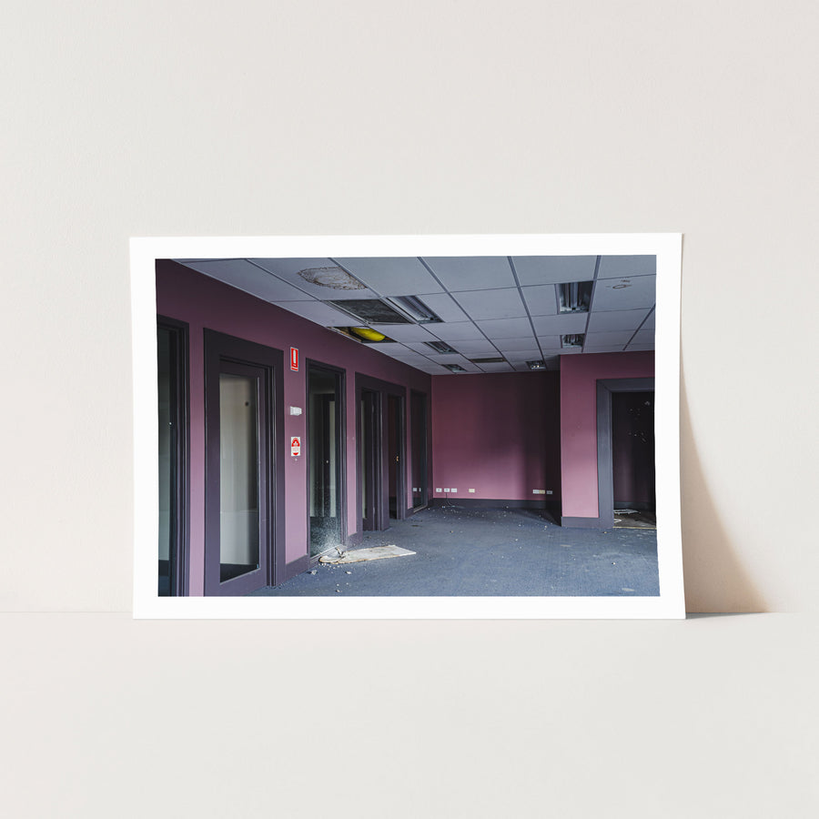 Empty office corridor at the former Bradmill factory in Yarraville, with mauve walls, grey carpet, glazed timber-framed doorways, a damaged suspended ceiling, and a red fire safety sign on the wall.
