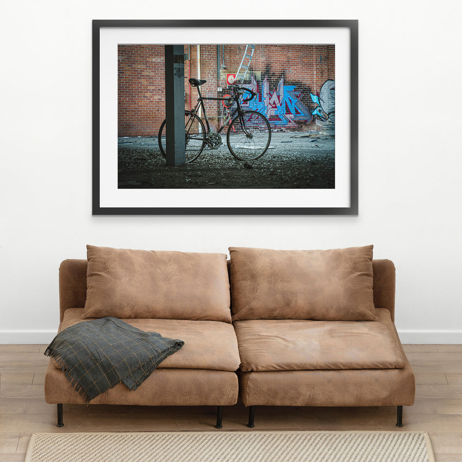 A worn black road bicycle leaning against a concrete pillar at the former Bradmill factory in Yarraville, with graffiti-covered brick wall, metal ladder, and red circular target visible behind it.