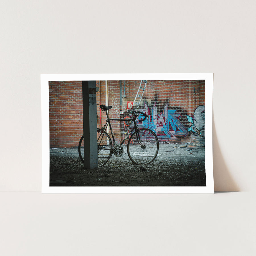 A worn black road bicycle leaning against a concrete pillar at the former Bradmill factory in Yarraville, with graffiti-covered brick wall, metal ladder, and red circular target visible behind it.