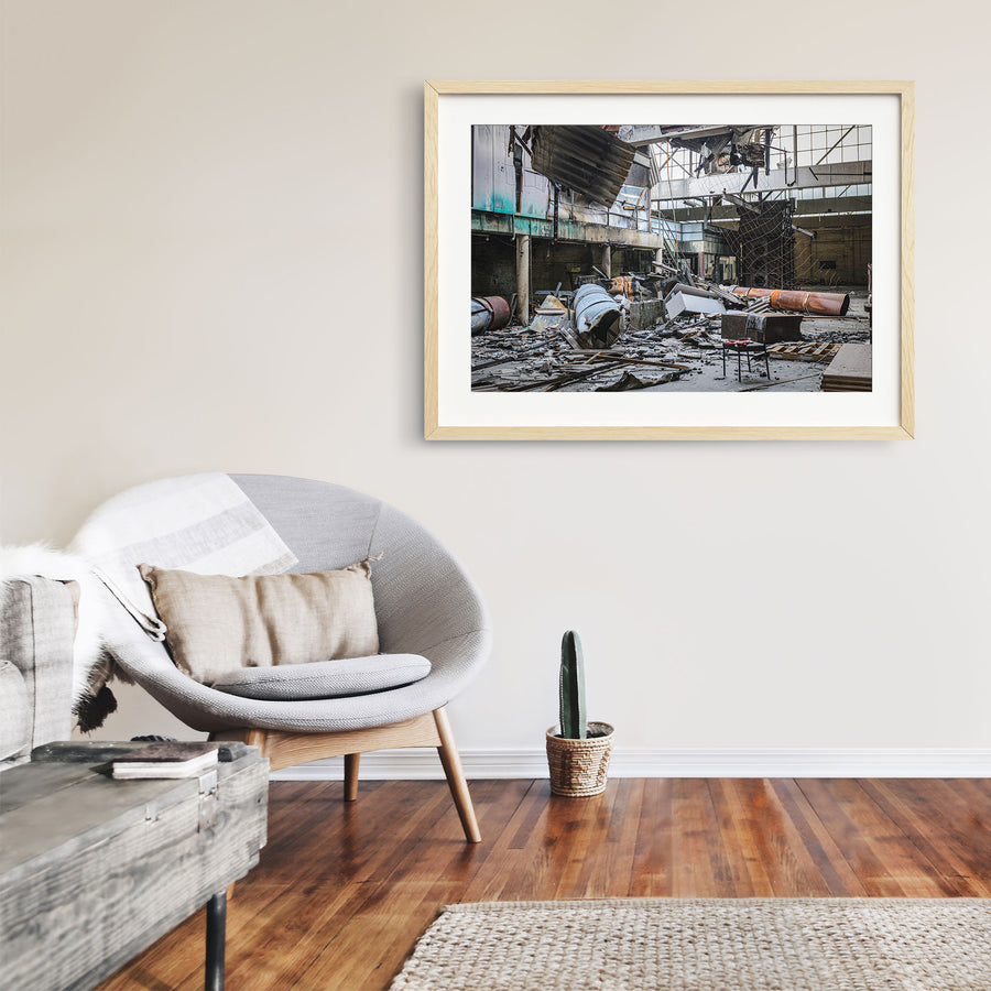 Fire-damaged manufacturing hall at the former Bradmill factory in Yarraville, with a collapsed roof, twisted metal sheeting, rusted cylindrical vessels, and a single chair on the debris-covered floor.