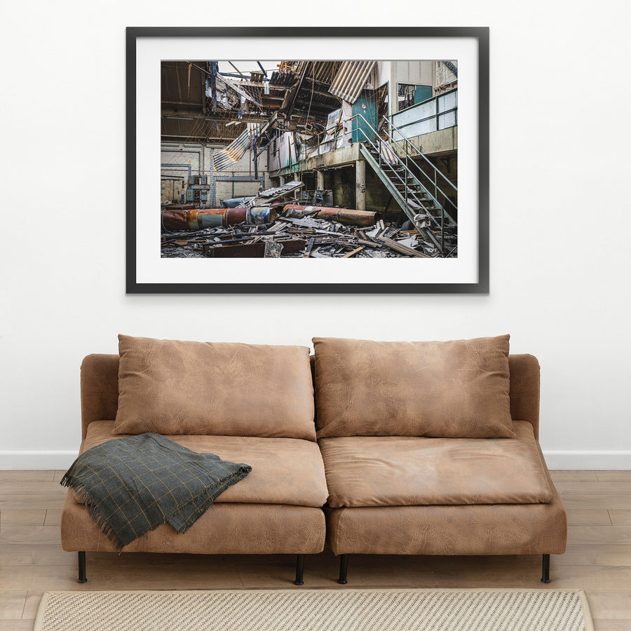 Collapsed corrugated roof sheeting, burnt timber and toppled cylindrical tanks covering the floor of a large industrial hall at the former Bradmill factory in Yarraville, with a steel staircase rising to a concrete mezzanine and old electrical boxes along a pale brick wall.