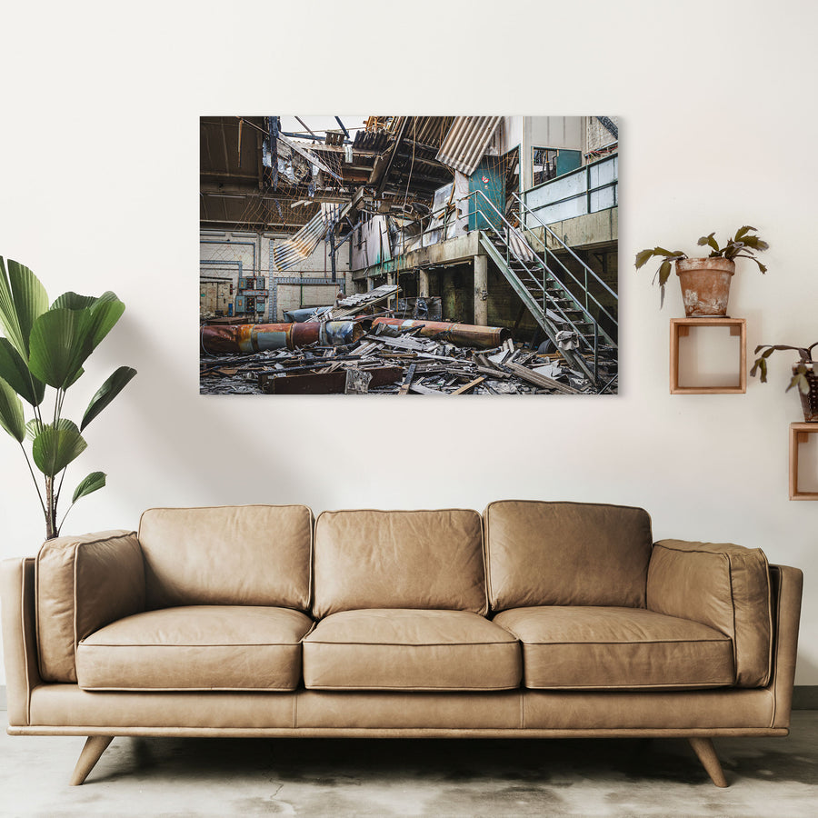 Collapsed corrugated roof sheeting, burnt timber and toppled cylindrical tanks covering the floor of a large industrial hall at the former Bradmill factory in Yarraville, with a steel staircase rising to a concrete mezzanine and old electrical boxes along a pale brick wall.