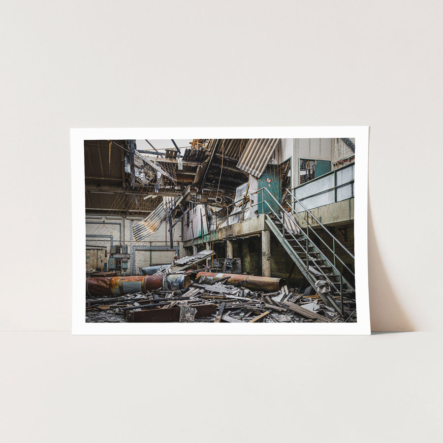 Collapsed corrugated roof sheeting, burnt timber and toppled cylindrical tanks covering the floor of a large industrial hall at the former Bradmill factory in Yarraville, with a steel staircase rising to a concrete mezzanine and old electrical boxes along a pale brick wall.