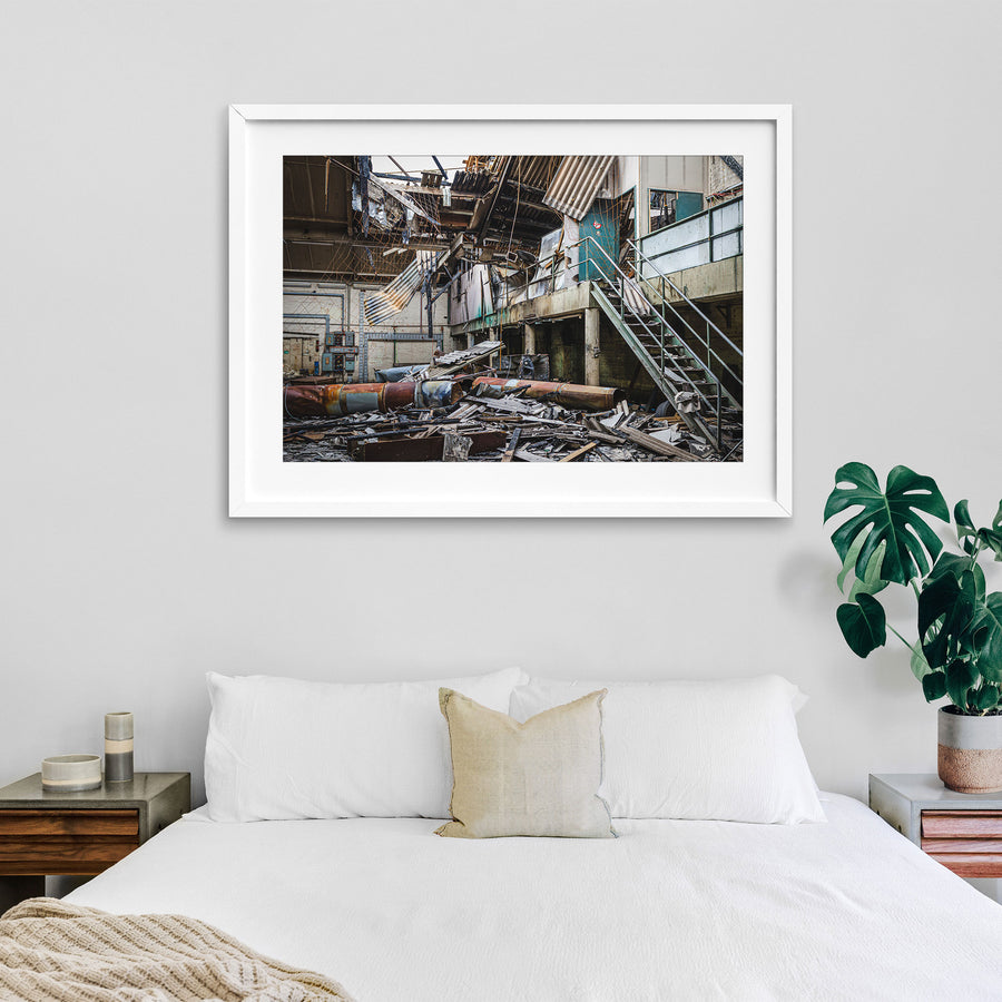 Collapsed corrugated roof sheeting, burnt timber and toppled cylindrical tanks covering the floor of a large industrial hall at the former Bradmill factory in Yarraville, with a steel staircase rising to a concrete mezzanine and old electrical boxes along a pale brick wall.