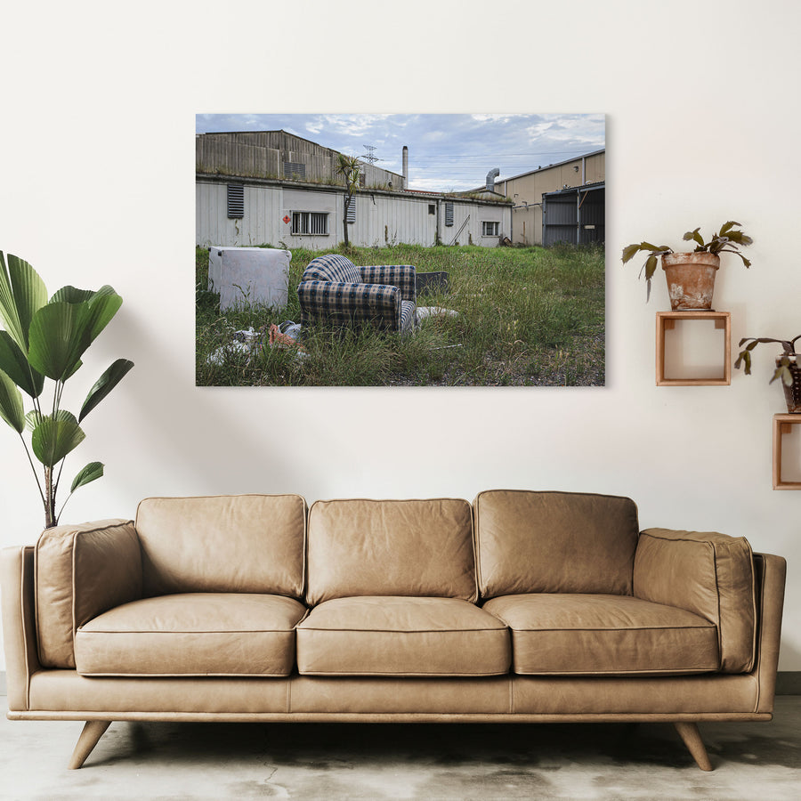 A patterned armchair sitting in long grass in front of metal-clad industrial sheds and a tall chimney at the former Davies Coop / Bradmill factory, Yarraville.