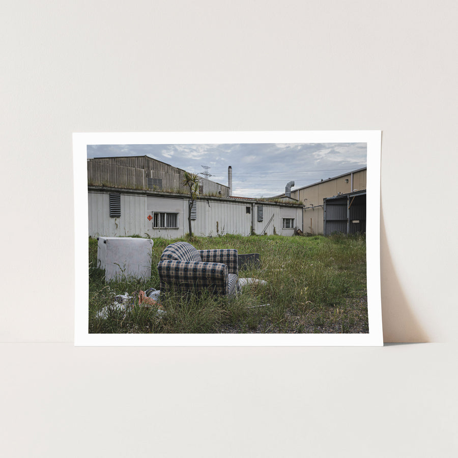 A patterned armchair sitting in long grass in front of metal-clad industrial sheds and a tall chimney at the former Davies Coop / Bradmill factory, Yarraville.