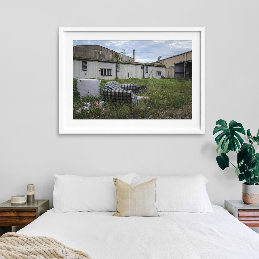 A patterned armchair sitting in long grass in front of metal-clad industrial sheds and a tall chimney at the former Davies Coop / Bradmill factory, Yarraville.