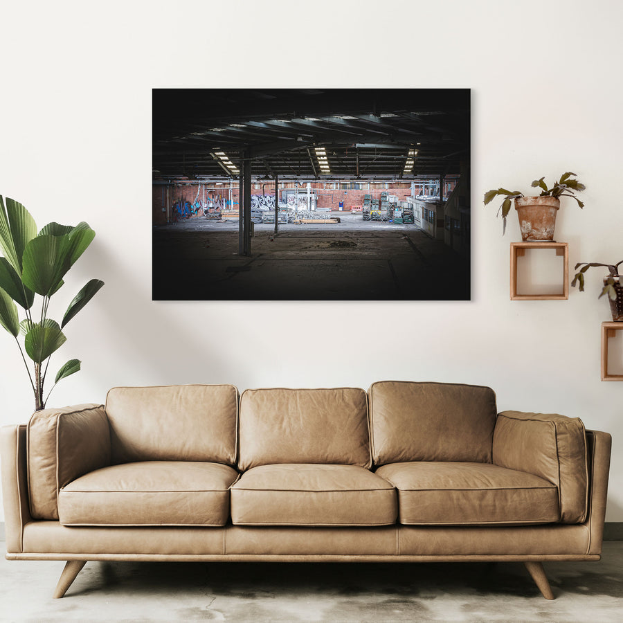 Empty warehouse interior at the former Bradmill factory in Yarraville, showing a steel-framed roof with skylights, graffiti-covered brick walls, and pallets and stacked timber across the concrete floor.