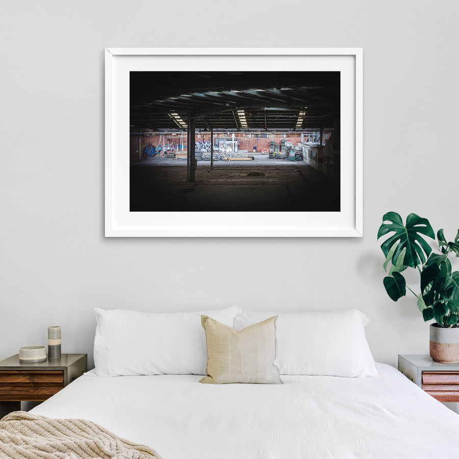 Empty warehouse interior at the former Bradmill factory in Yarraville, showing a steel-framed roof with skylights, graffiti-covered brick walls, and pallets and stacked timber across the concrete floor.