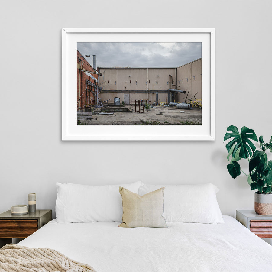 A concrete yard at the former Bradmill factory in Yarraville, enclosed by rendered walls and a brick building, with disused ducting, metal drums, a galvanised tub and severed pipework left across the slab under an overcast sky.