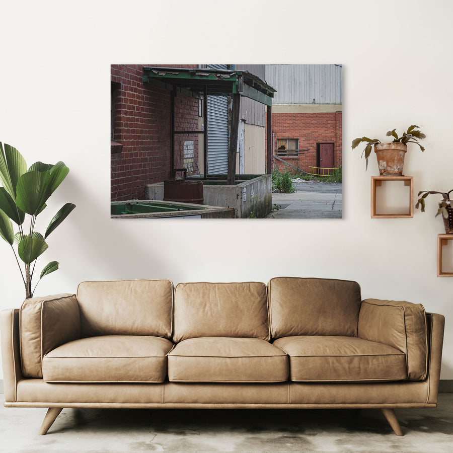 A green steel canopy over a concrete loading dock at the former Bradmill factory in Yarraville, with a closed corrugated roller door and a rusted metal bin below, a cracked yard and brick warehouse wall with a broken window beyond.