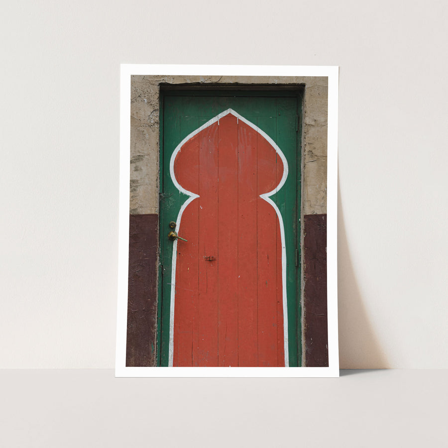 A painted timber door at the former Bradmill factory in Yarraville, with a tall red keyhole arch outlined in white on a green background, a worn brass lever handle on the left, and cracked render surrounding the frame.