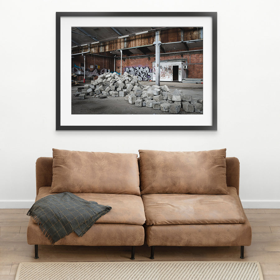 A pile of grey stone blocks on the dusty floor of an abandoned brick manufacturing building at the former Bradmill factory, Yarraville, with graffiti-covered walls and a rusting roof beam above.