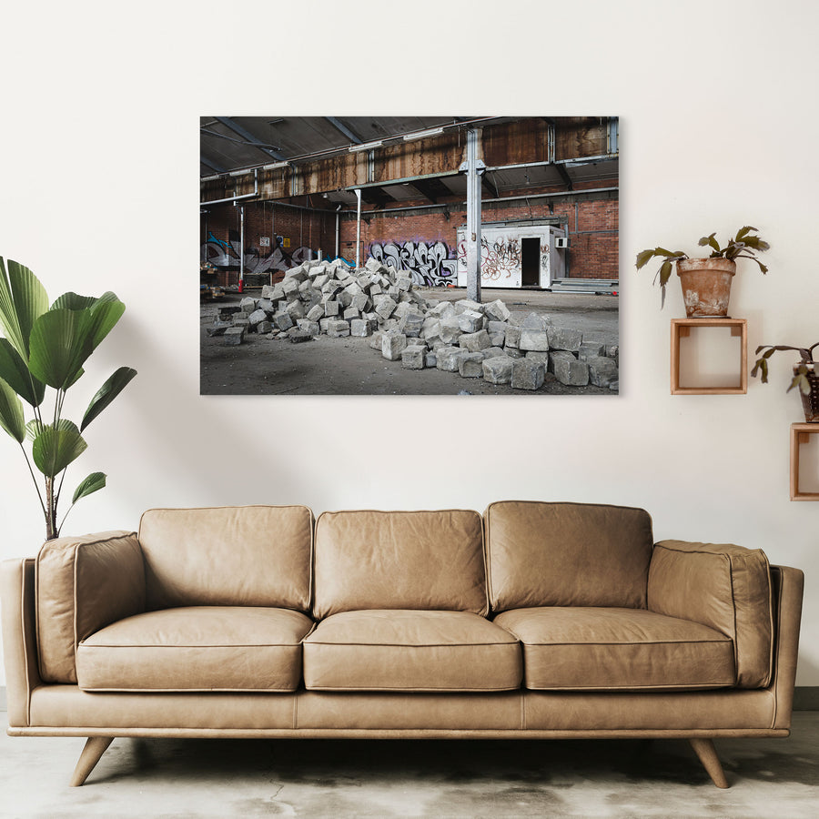 A pile of grey stone blocks on the dusty floor of an abandoned brick manufacturing building at the former Bradmill factory, Yarraville, with graffiti-covered walls and a rusting roof beam above.