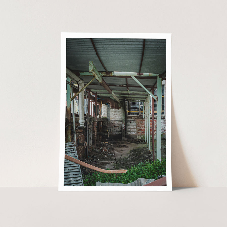 Rusting steel-framed canopy over a derelict brick interior at the former Davies Coop / Bradmill factory in Yarraville, with exposed pipework, a coiled hose and weeds encroaching on the concrete floor.