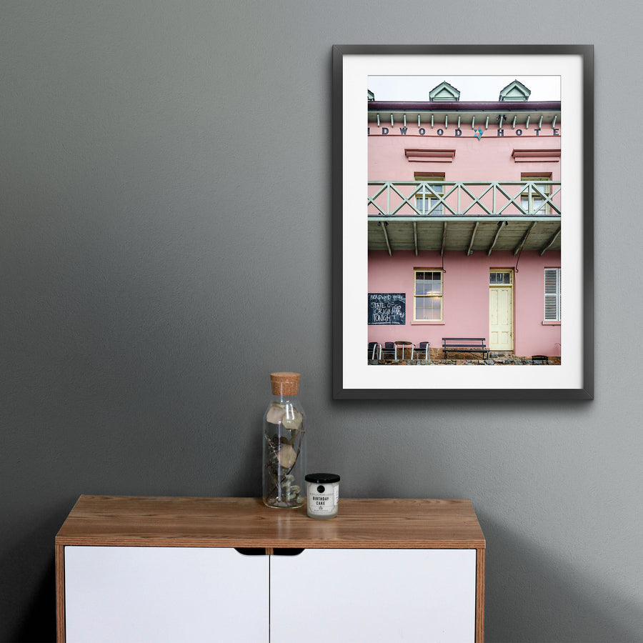 The pink rendered two-storey facade of Braidwood Hotel, with a timber balcony above a verandah, a chalkboard sign, and outdoor seating at street level.