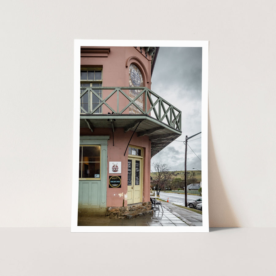 Pink rendered two-storey hotel on a corner footpath at Braidwood, with green timber balcony railings, yellow bar doors, beer signage, and metal tables on wet pavement under an overcast sky.