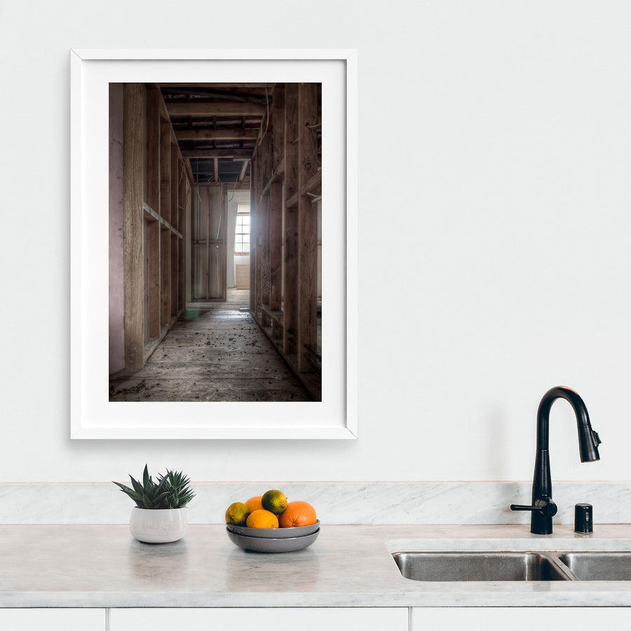 Exposed timber wall framing and ceiling joists in a stripped corridor at the Braidwood Hotel, with bare floorboards leading to a multi-pane window partially covered by plywood.
