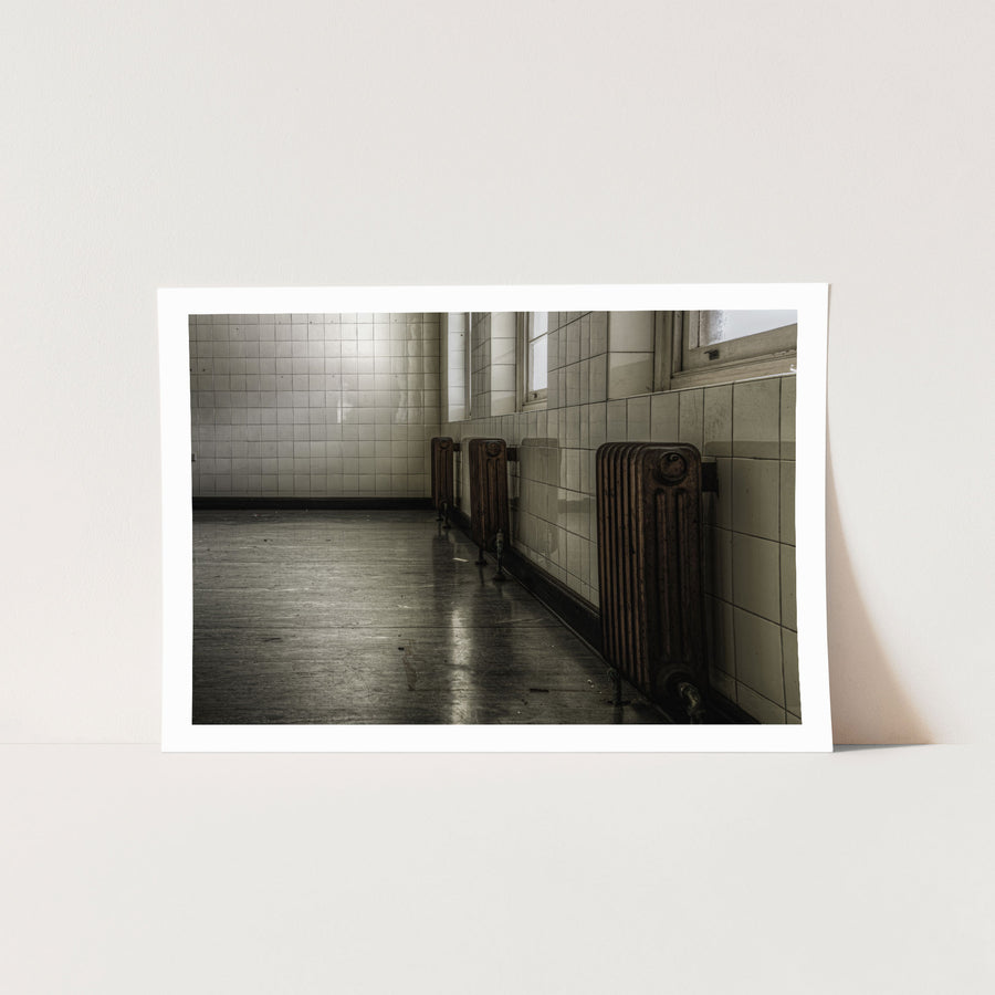 Three cast-iron radiators along a tiled corridor wall at Callan Park, reflected in the polished concrete floor below.