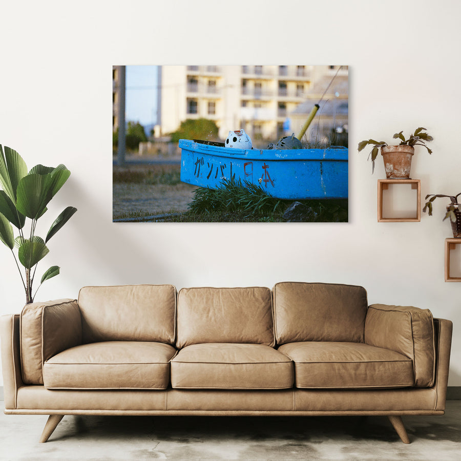 A weathered blue boat with ガンバレ日本 painted on its hull rests on dry grass at Ikeshima, a white ceramic figure inside and a concrete apartment block behind.