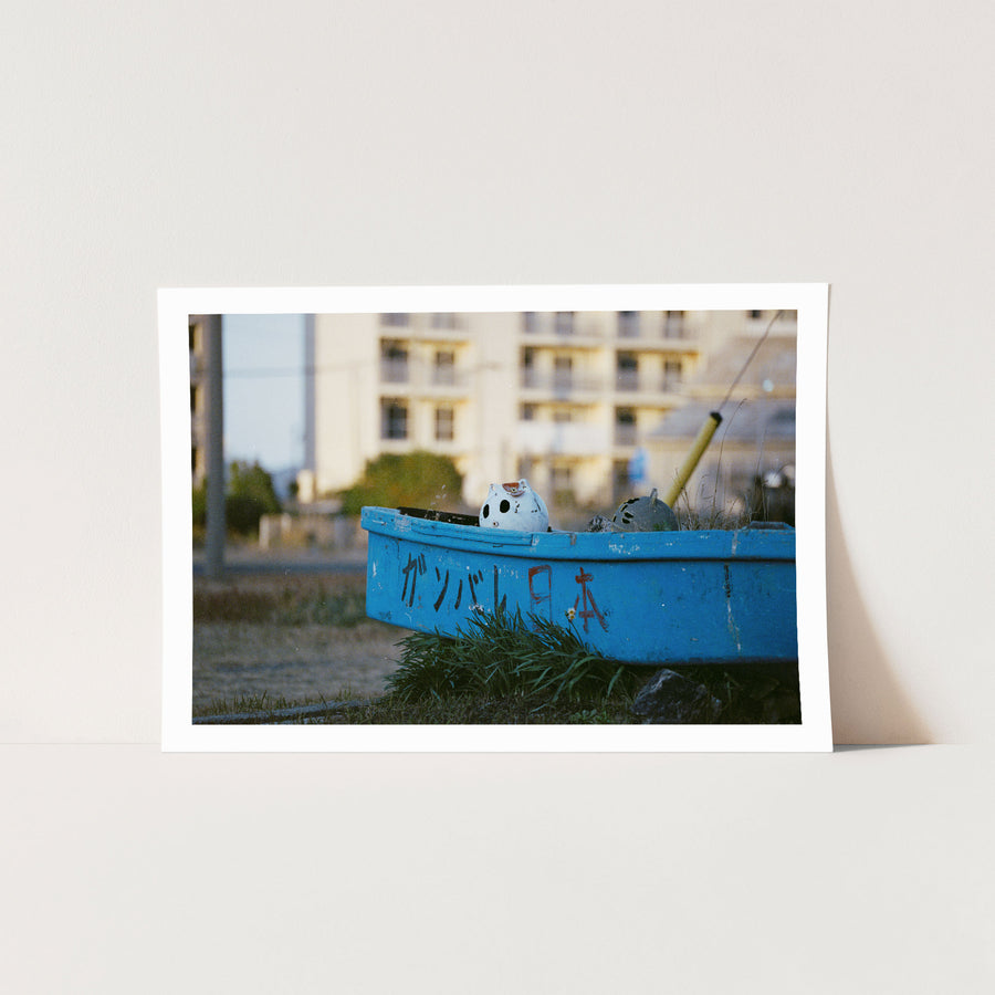 A weathered blue boat with ガンバレ日本 painted on its hull rests on dry grass at Ikeshima, a white ceramic figure inside and a concrete apartment block behind.