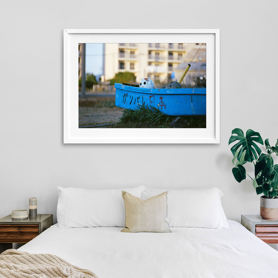 A weathered blue boat with ガンバレ日本 painted on its hull rests on dry grass at Ikeshima, a white ceramic figure inside and a concrete apartment block behind.