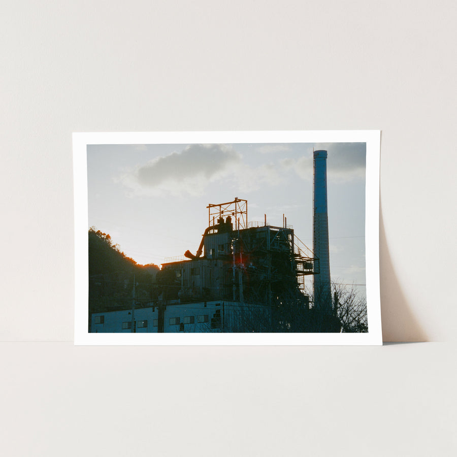 The chimney and boiler house of the coal-fired power station at Ikeshima Coal Mine silhouetted against a hazy sunset sky, with external pipework, steel walkways and a forested hillside visible in the frame.