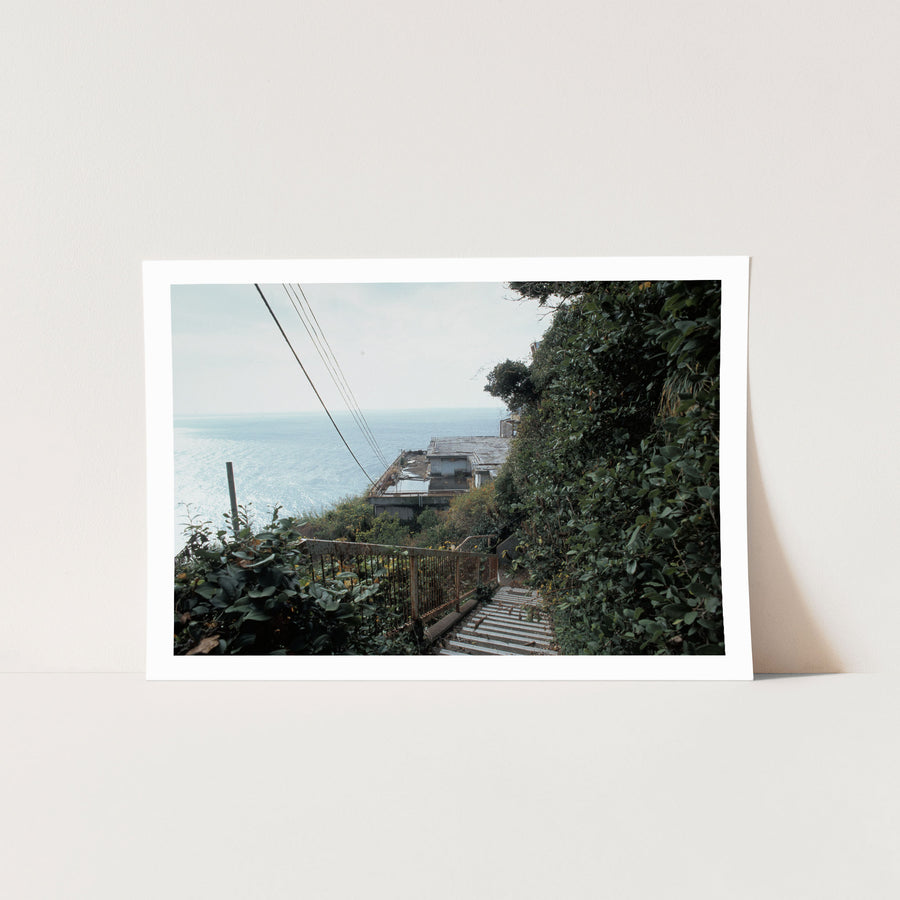 A steep concrete staircase with a rusted railing descends through dense green foliage at Ikeshima, with power lines crossing a bright sky and the sea visible below.