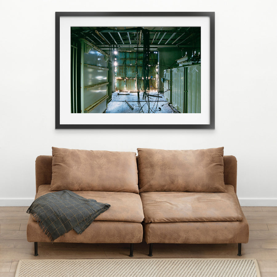 Dense black cable bundle hanging from an exposed ceiling frame runs the length of a corridor at Ikeshima Coal Mine, with two ribbed transformers along one wall and teal double doors at the far end.