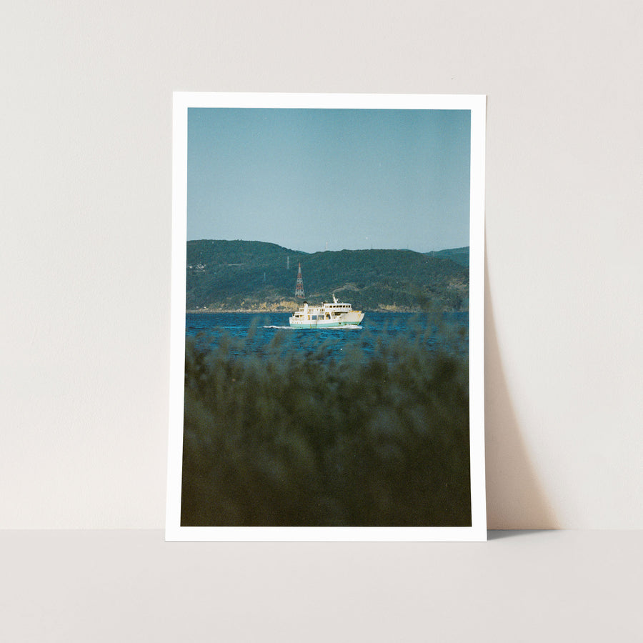 A white and teal ferry crosses the channel at Ikeshima, trailing a bright wake, with a red and white lattice tower rising above the forested hillside behind the port.