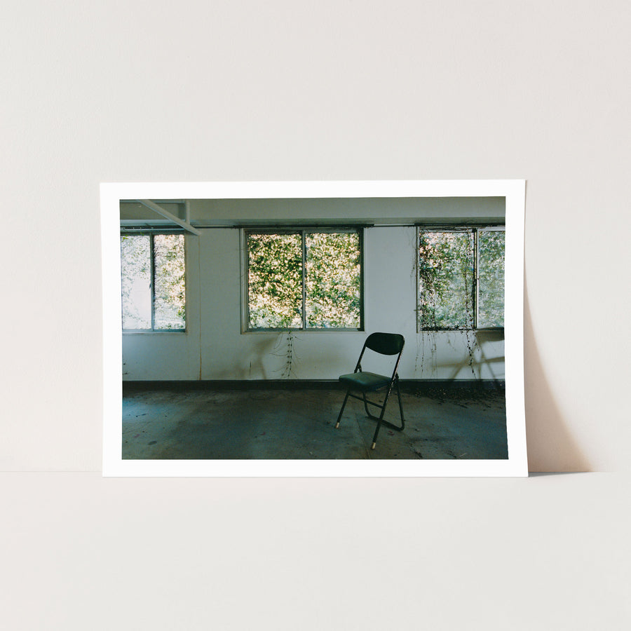 A dark green folding chair on a dusty concrete floor faces three large windows thick with green foliage, with vine tendrils trailing down the white wall beside water stains, in an office at the Ikeshima Coal Mine.