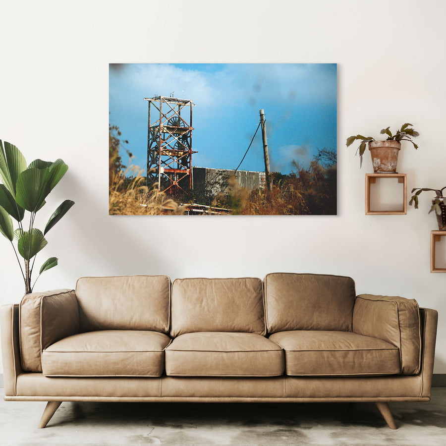 A rusting steel lattice headframe with two spoked sheave wheels silhouetted against a bright sky at Ikeshima Coal Mine, with dry grasses blurring across the foreground.