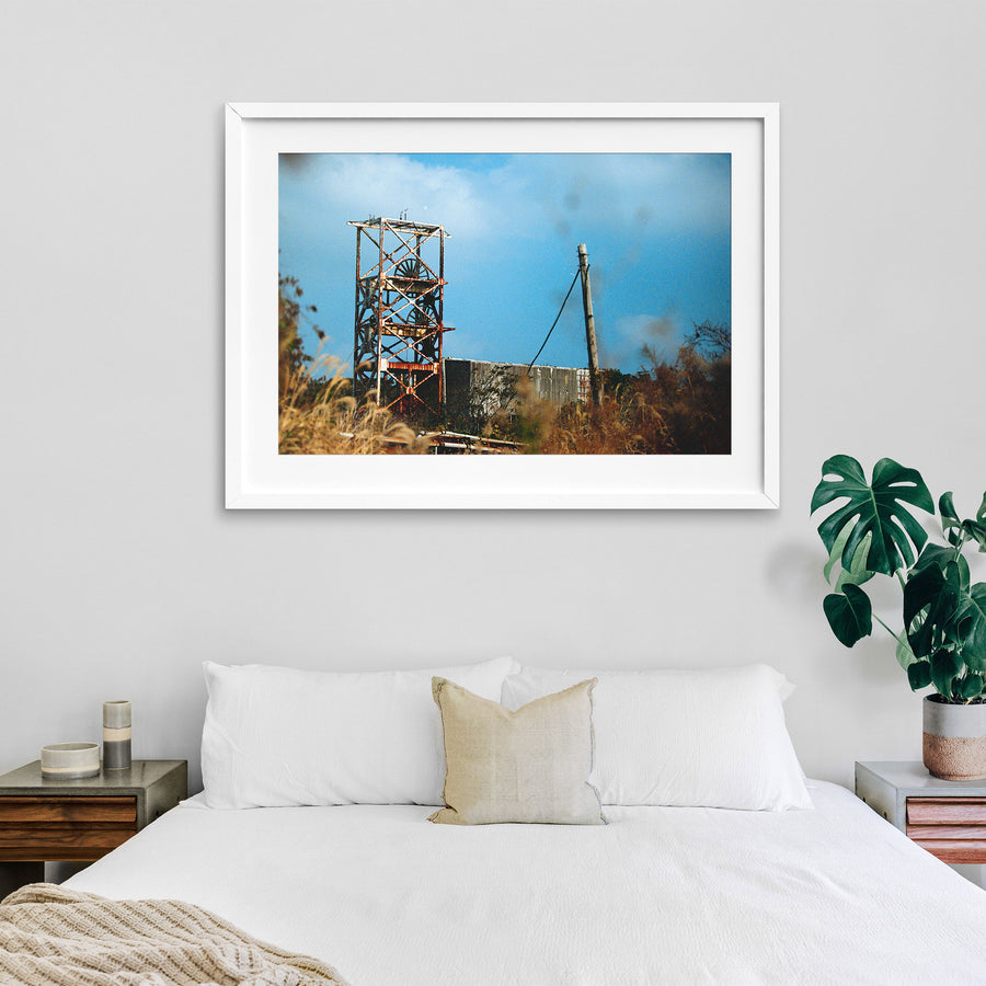 A rusting steel lattice headframe with two spoked sheave wheels silhouetted against a bright sky at Ikeshima Coal Mine, with dry grasses blurring across the foreground.