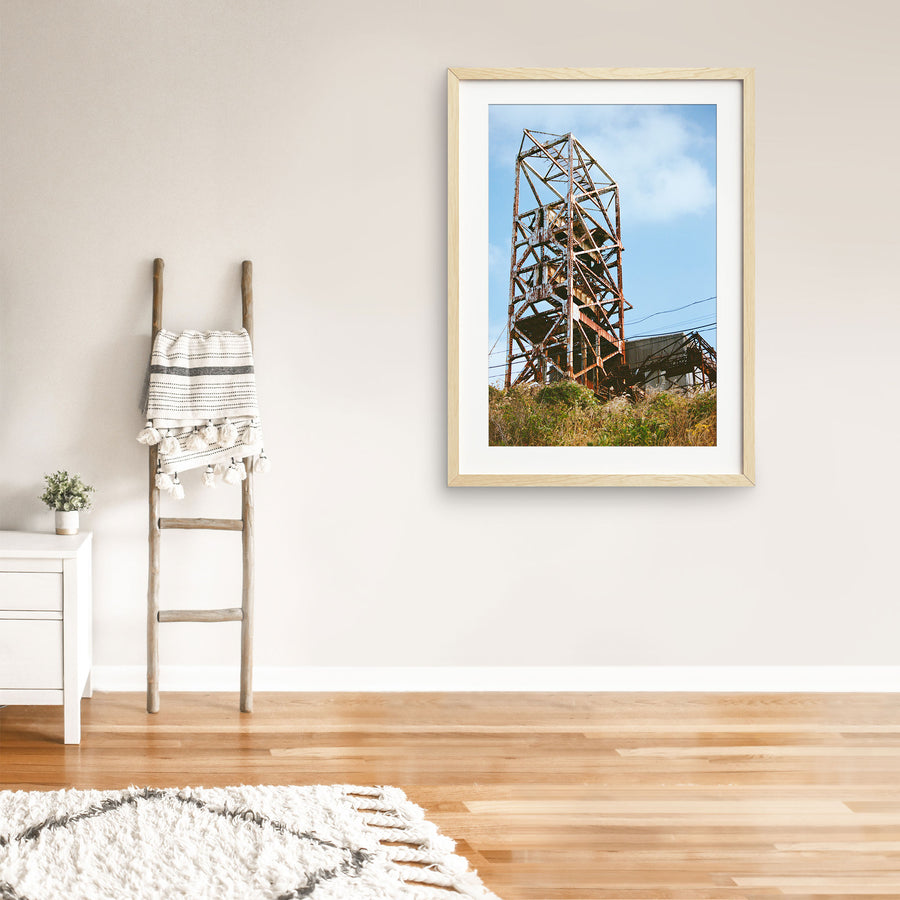 Rusted steel lattice of a mine headframe at Ikeshima, with power lines crossing toward a corrugated shed and dry golden grass on the slope below.
