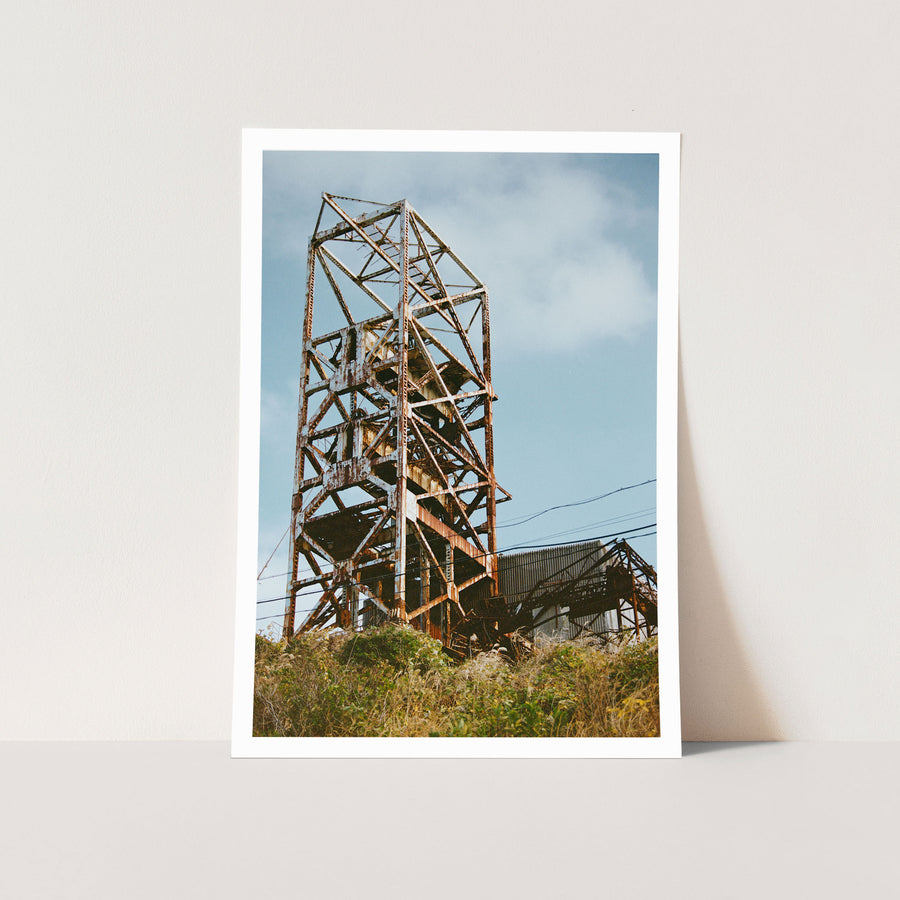 Rusted steel lattice of a mine headframe at Ikeshima, with power lines crossing toward a corrugated shed and dry golden grass on the slope below.